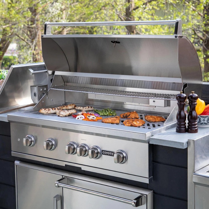 Stainless steel outdoor grill with an open lid, featuring various meats and vegetables cooking on the grates, alongside condiments and spices on a countertop.