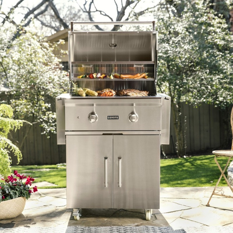 Stainless steel outdoor grill with an open lid displaying grilled meats and vegetables, set in a backyard with greenery and flowers.