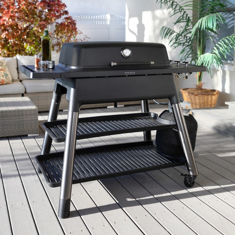 Black outdoor grill with a temperature gauge, side shelves, and a propane tank, set on a wooden deck with patio furniture in the background.