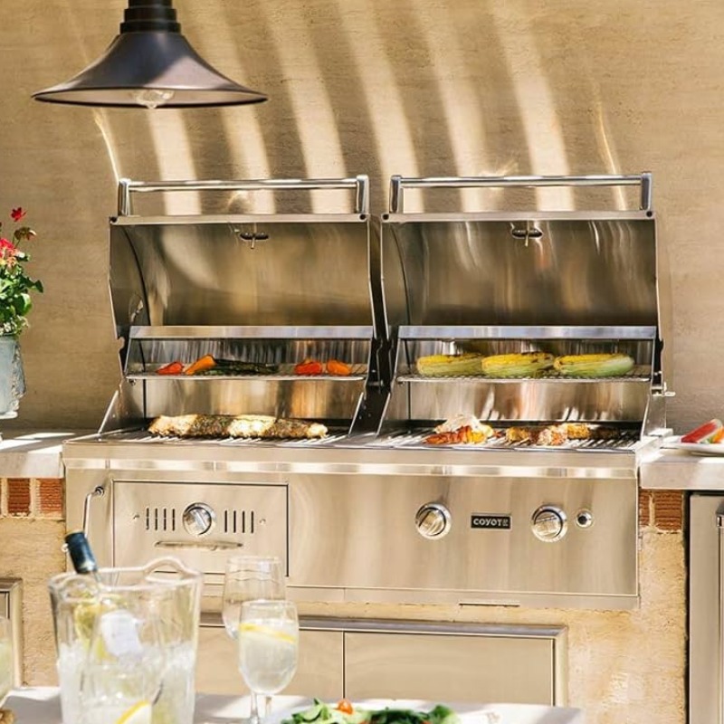 Stainless steel outdoor grill with two cooking compartments, featuring various grilled vegetables and meats, set in a sunny patio with drinks and greenery.