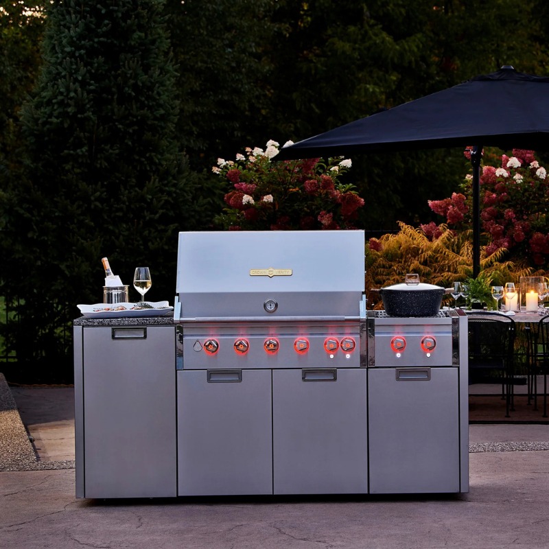Stainless steel outdoor grill with multiple burners, granite countertop, and cooking accessories, set in a landscaped garden with flowers and outdoor seating.