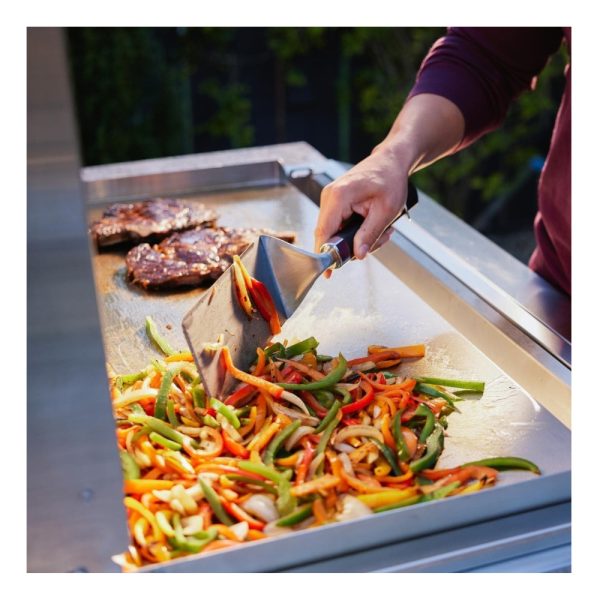A person using a spatula to stir-fry colorful bell peppers and onions on a grill, with grilled steaks in the background.
