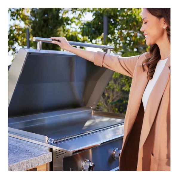 A woman in a beige coat opens the lid of a modern stainless steel grill in an outdoor setting, smiling as she prepares to cook.