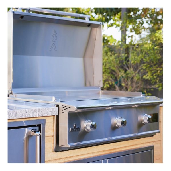 Stainless steel outdoor grill with three knobs and a raised lid, set against a backdrop of greenery.