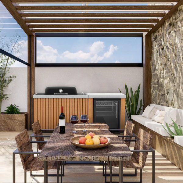 Outdoor kitchen with wooden pergola, modern grill, and dining table set. Features include a wine bottle, glasses, and a fruit bowl, surrounded by plants and stone walls. Ideal for entertaining.